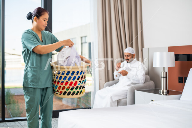 رجل عربي سعودي خليجي يجلس على الريكة ويشرب القهوة، عاملة فلبينية ترتدي اليونيفورم الخاص بالعمل تحمل سلة غسيل مليئة بالملابس، الاهتمام بالمهام والأعمال المنزلية الروتينية، استقدام العمالة الخارجية المرخصة والمؤهلة