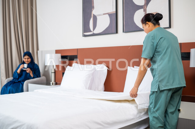 استقدام العمالة الخارجية المرخصة والمؤهلة، امرأة عربية سعودية خليجية تجلس على الاريكة وتشرب القهوة، عاملة فلبينية ترتدي اليونيفورم الخاص بالعمل تقوم بترتيب السرير في غرفة النوم، الاهتمام بالمهام والأعمال المنزلية الروتينية