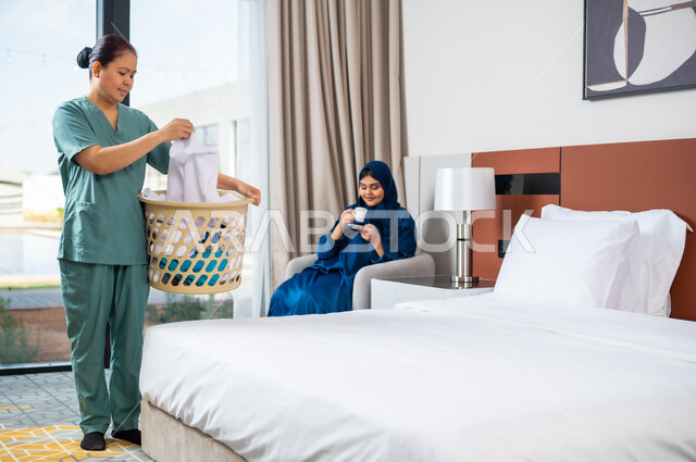 الاهتمام بالمهام والأعمال المنزلية الروتينية، استقدام العمالة الخارجية المرخصة والمؤهلة، امرأة عربية سعودية خليجية تجلس على الاريكة وتشرب القهوة، عاملة فلبينية ترتدي اليونيفورم الخاص بالعمل تقوم بجمع الملابس في سلة الغسيل
