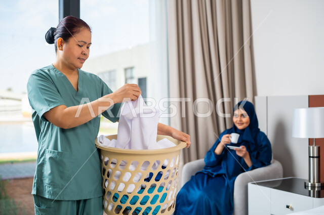 Recruiting licensed and qualified foreign workers, a Saudi Arabian Gulf woman sitting on the sofa and drinking coffee, a Filipina worker wearing her work uniform, collecting clothes in the laundry basket, taking care of routine tasks and household chores.