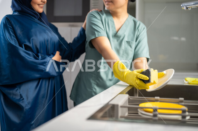 استقطاب العمالة الأجنبية المرخصة والمؤهلة من الخارج، سيدة عربية خليجية سعودية تقوم باعطاء التعليمات للخادمة بايماءات الرضا, التطهير والتنظيف وغسل المواعين, أداء مهام المطبخ، عاملة أجنبية ترتدي اليونيفورم الخاص والقفازات تقوم بغسيل الأطباق باستخدام اسفنجة الجلي