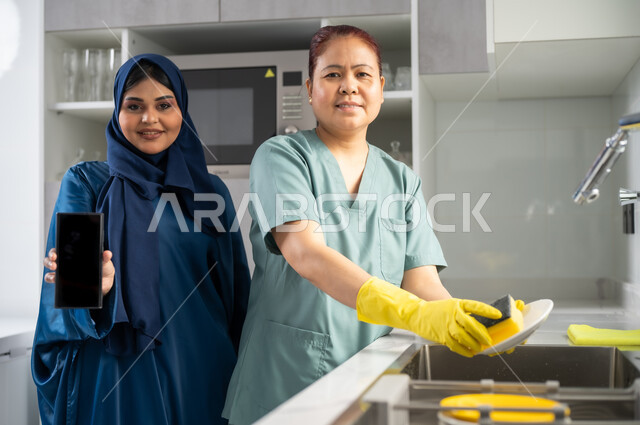 أداء مهام المطبخ, عاملة أجنبية ترتدي اليونيفورم الخاص والقفازات تقوم بغسيل الأطباق باستخدام اسفنجة الجلي, استقطاب العمالة الأجنبية المرخصة والمؤهلة من الخارج, التطهير والتنظيف وغسل المواعين،امرأة عربية سعودية خليجية تقوم بعرض شاشة سوداء فارغة على الهاتف المحمول