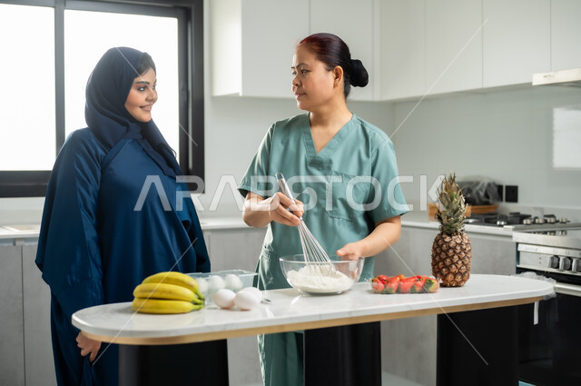 تشغيل العاملات المرخصة والمؤهلة من الشركات والمكاتب السعوديه, تحضير وتجهيز المكونات اللازمة, اعداد الحلويات الشهية في المطبخ, عاملة أجنبية ترتدي اليونيفورم الخاص تقوم بصنع الحلى برفقة ربة المنزل
