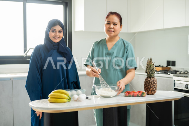 تشغيل العاملات المرخصة والمؤهلة من الشركات والمكاتب السعوديه, تحضير وتجهيز المكونات اللازمة, اعداد الحلويات الشهية في المطبخ, عاملة أجنبية ترتدي اليونيفورم الخاص تقوم بصنع الحلى برفقة ربة المنزل, النظر الى الكاميرا بايماءات السعاة والرضا