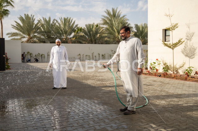 التسلية والترفيه وقضاء الوقت الممتع مع الأصدقاء في إحدى الشاليهات, مفهوم النظافة والاهتمام بحديقة المنزل, رجل عربي خليجي سعودي يرتدي الثوب التقليدي يقوم برش الحديقة بالمياه, أماكن سياحية ترفيهية