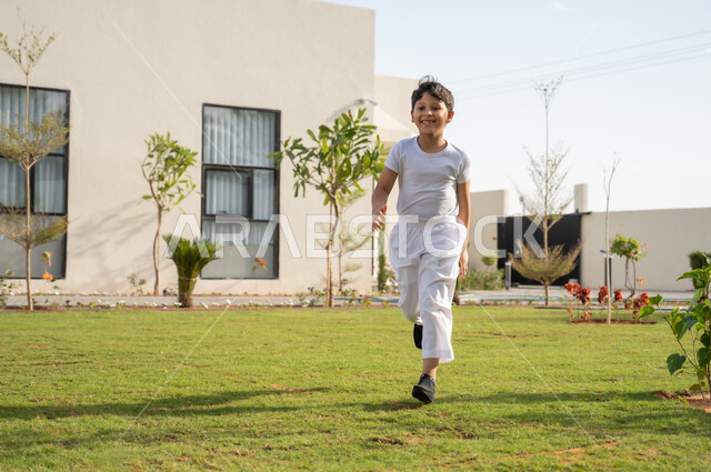 اللعب في حديقة المنزل بإيماءات السعادة والسرور, التسلية والترفيه وقضاء الوقت الممتع, فتى عربي خليجي سعودي يرتدي السروال والفانيله يركض في الحديقة الخارجية, نزهة في الهواء الطلق بين الأشجار والنباتات الخضراء, الركض في إحدى الشاليهات والمنتجعات السياحية الراقية
