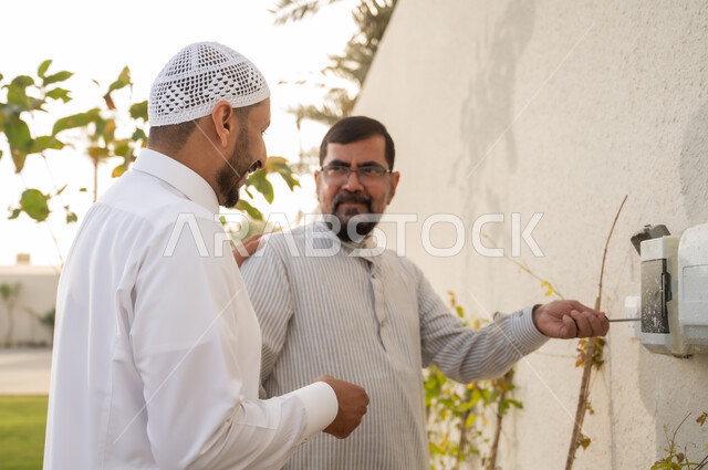 محاولة فتح علبة الكهرباء، صورة مقربة من الجانب لرجلان عربيان خليجيان سعوديان يرتديان الثوب التقليدي يقومان باستخدام مفك الفحص, اصلاح وصيانة الأعطال الكهربائية في حديقة المنزل, استخدام الالات والمعدات اليدوية