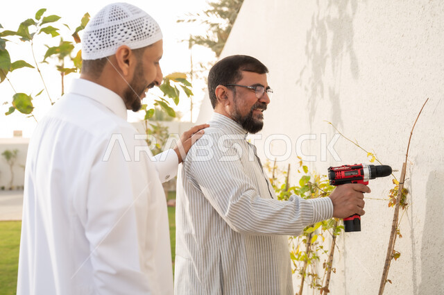 معدات الحفر والتثقيب، حرف ومهن يدوية، أدوات الإصلاح والصيانة الكهربائية, رجلان عربيان خليجيان سعوديان يرتديان الثوب التقليدي يقومان باستخدام المثقب الكهربائي في حديقة المنزل، إنجازمهام العمل