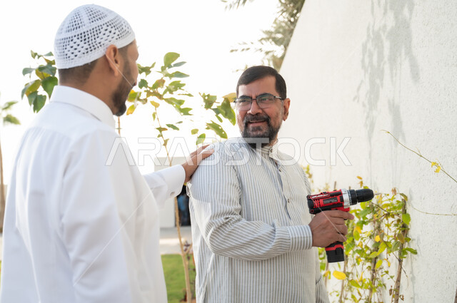 أدوات الإصلاح والصيانة الكهربائية, إنجازمهام العمل، رجلان عربيان خليجيان سعوديان يرتديان الثوب التقليدي يقومان باستخدام المثقب الكهربائي في حديقة المنزل, معدات الحفر والتثقيب، حرف ومهن يدوية