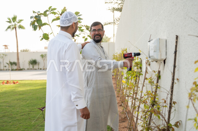 إنجازمهام العمل، رجلان عربيان خليجيان سعوديان يرتديان الثوب التقليدي يقومان باستخدام المثقب الكهربائي في حديقة المنزل, أدوات الإصلاح والصيانة الكهربائية, معدات الحفر والتثقيب، حرف ومهن يدوية