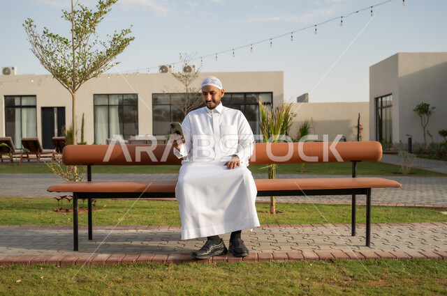 تصفح وسائل التواصل الاجتماعي, دردشات ومحادثات مع الأهل والاصدقاء عبر الجوال, رجل عربي خليجي سعودي يرتدي الثوب التقليدي يمسك بيده الهاتف المحمول, الاستمتاع بالجلسات والأجواء الهادئة في حديقة المنزل