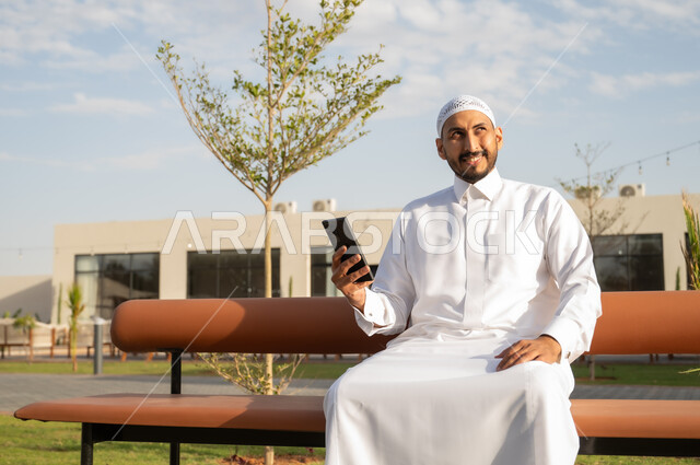 اجراء مكالمة فيديو عبر الهاتف المحمول, استخدام الأجهزة والتقنيات الحديثة والمتطورة, الجلسات والأجواء الهادئة في النهار, التسلية والترفيه عبر السوشال ميديا, رجل عربي خليجي سعودي يرتدي الثوب التقليدي يقوم باستخدام الجوال وينظر الى الكاميرا