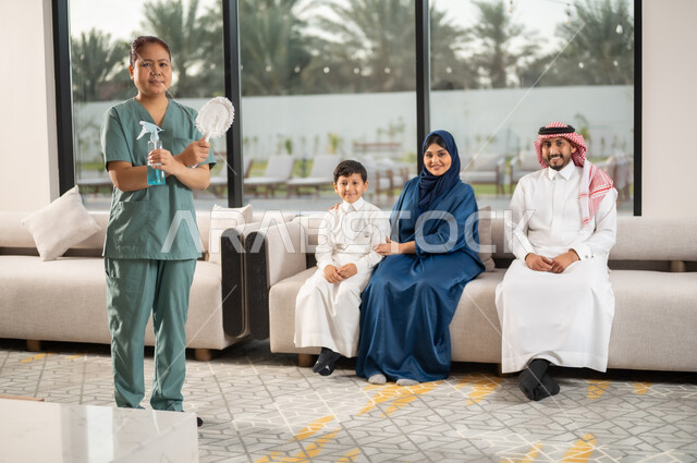توفير خدمات العناية بالمنزل والنظافة والترتيب، التنظيف والاهتمام بالأعمال الروتينية اليومية, استقطاب عاملات أجنبيات مرخصة ومؤهلة من الخارج, عاملة أجنبية ترتدي اليونيفورم الخاص تحمل ادوات التنظيف تنظر الى الكاميرا عائلة بايماءات السرور