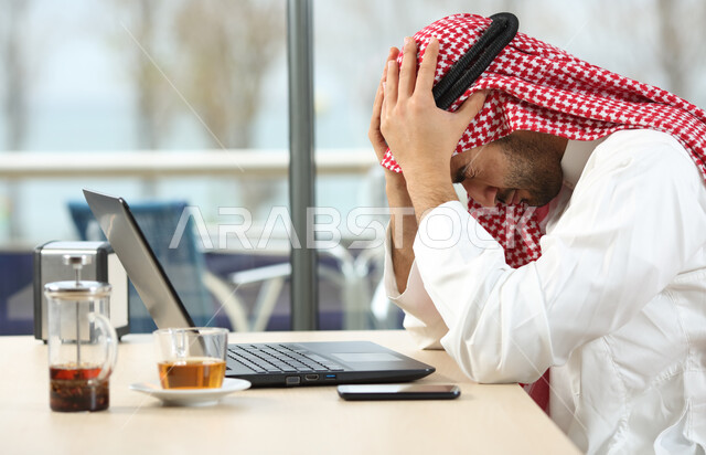 مفهوم ضغط العمل , مهن و وظائف مكتبية في السعودية , رجل أعمال عربي خليجي سعودي يرتدي الثوب التقليدي و الشماغ يجلس على الطاولة أمام جهاز الحاسوب و يضع يديه على رأسه بإيماءات التوتر و الانزعاج , تنظيم و إدارة الأعمال ضمن مقر الشركة