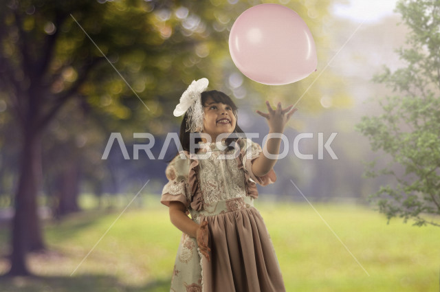 اجواء احتفالية بمناسبة قدوم العيد  لفتاه صغيرة سعودية خليجية مبتسمة وجميلة ، تلعب بالبالون زهري اللون بإيماءات وجه ويدين مختلفة تدل على السعادة ,  ترتدي لباس العيد الانيق , اعياد و مناسبات