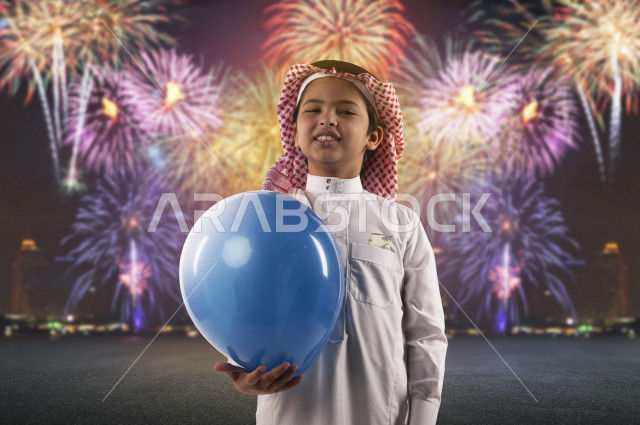 صبي صغير سعودي خليجي مبتسم وجميل ، يلعب بالبالون أزرق اللون بإيماءات وجه ويدين مختلفة تدل على السعادة , يرتدي الزي السعودي التقليدي , الاعياد و المناسبات ، خلفية لسماء مليئة  بالالعاب النارية