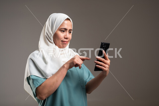 الدردشة مع الأصدقاء والاقارب، مواكبة التطور التقني والتقدم التكنولوجي، استخدام الأجهزة التقنية الحديثة والمتطورة، بورتريه لعاملة فلبينية تحمل هاتف محمول،خلفية رمادية