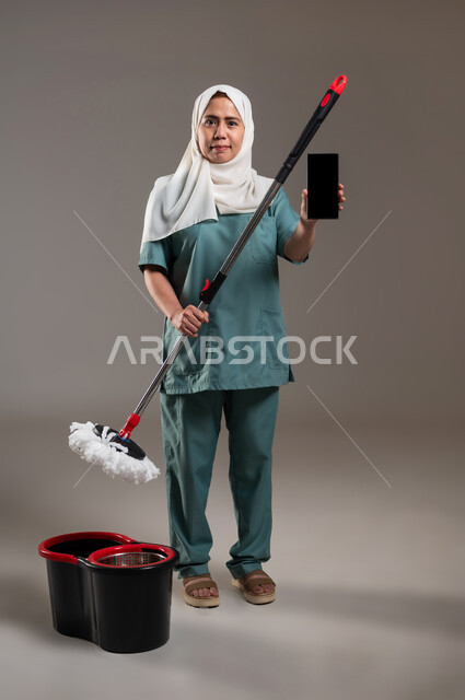 دور الهاتف المحمول في حياتنا، القيام بالأعمال المنزلية، التكنولوجيا والتطور التقني،استخدام الادوات الكهربائية لإنجاز العمل، مكنسة بلون أحمر واسود، بورتريه لعاملة فلبينية محجبة ترتدي اليونيفورم تقوم بأعمال التنظيف، الطول الكامل للجسم، خلفية رمادية