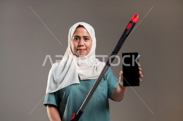 دور الهاتف المحمول في حياتنا، القيام بالأعمال المنزلية، التكنولوجيا والتطور التقني،استخدام الادوات الكهربائية لإنجاز العمل، مكنسة بلون أحمر واسود، بورتريه لعاملة فلبينية محجبة ترتدي اليونيفورم تقوم بأعمال التنظيف، خلفية رمادية