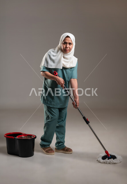 الاستعداد لبدء العمل، القيام بالأعمال المنزلية، أدوات تنظيف بلون أحمر واسود، بورتريه لعاملة فلبينية محجبة ترتدي اليونيفورم تقوم بأعمال التنظيف، الطول الكامل للجسم، خلفية رمادية