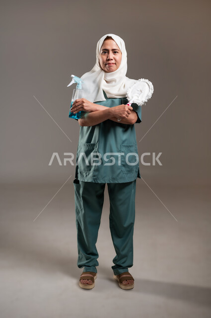ممسحة وملمع اسطح، الاهتمام بنظافة وتطهير البيت والقيام بالمهام الروتينية, مهن ووظائف نسائية، بورتريه لعاملة منزلية فلبينية محجبة تحمل بيدها أدوات تنظيف المنزل، الجسم بالطول الكامل، خلفية رمادية