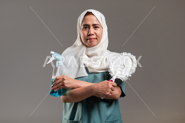 ممسحة وملمع اسطح، الاهتمام بنظافة وتطهير البيت والقيام بالمهام الروتينية, مهن ووظائف نسائية، بورتريه لعاملة منزلية فلبينية محجبة تحمل بيدها أدوات تنظيف المنزل، خلفية رمادية