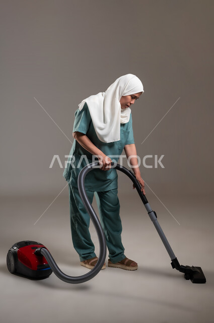 الاستعداد للقيام بالأعمال المنزلية، بورتريه لعاملة فلبينية محجبة ترتدي اليونيفورم الخاص بالعمل تقوم باستخدام الأدوات الكهربائية لإنجاز المهام الروتينية، الطول الكامل للجسم، استقدام العماله من الخارج، خلفية رمادية