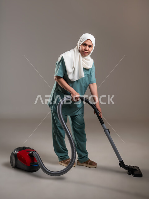 Preparing to do housework, portrait of veiled Filipina worker wearing work uniform using power tools to complete routine tasks, full body length, recruiting workers from abroad, gray background