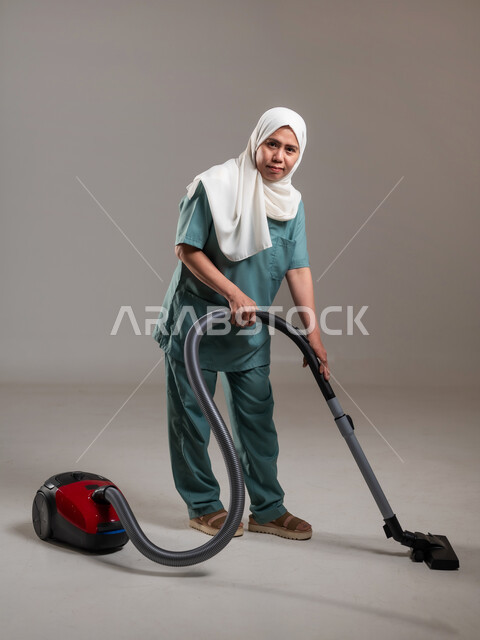 Preparing to do housework, portrait of veiled Filipina worker wearing work uniform using power tools to complete routine tasks, full body length, recruiting workers from abroad, gray background