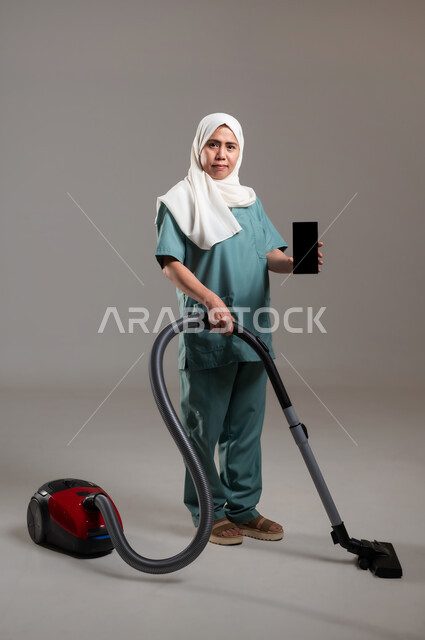 دور الهاتف المحمول في حياتنا، القيام بالأعمال المنزلية، التكنولوجيا والتطور التقني،استخدام الادوات الكهربائية لإنجاز العمل، مكنسة بلون أحمر واسود، بورتريه لعاملة فلبينية محجبة ترتدي اليونيفورم تقوم بأعمال التنظيف، الطول الكامل للجسم، خلفية رمادية