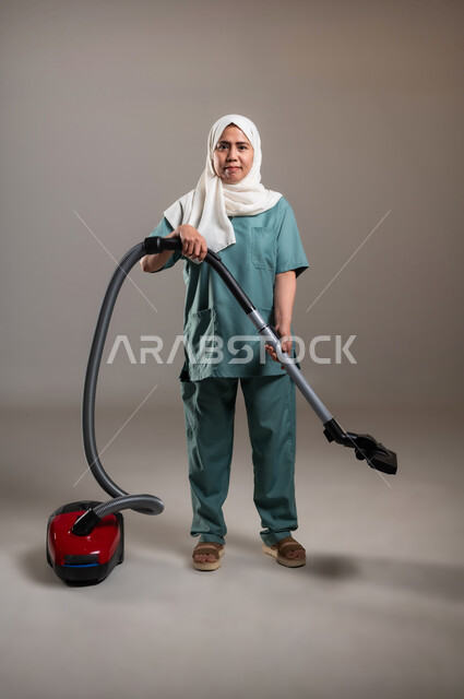 الاستعداد للقيام بالأعمال المنزلية، بورتريه لعاملة فلبينية محجبة ترتدي اليونيفورم الخاص بالعمل تقوم باستخدام الأدوات الكهربائية لإنجاز المهام الروتينية، الطول الكامل للجسم، استقدام العماله من الخارج، خلفية رمادية
