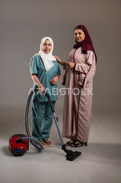 الاستعداد للقيام بالاعمال المنزلية الروتينية، النظر للكاميرا بتعابير السعادة والفرح، بورتريه لإمرأة عربية خليجية سعودية تقف إلى جانب مساعدة منزلية فلبينية ترتدي اليونيفورم الخاص بها تمسك بيدها مكنسة كهربائية، استقدام العماله من الخارج، الطول الكامل للجسم، خلفية رمادية