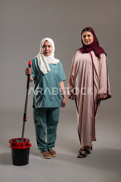 النظر للكاميرا بتعابير الابتسامة، بورتريه لإمرأة عربية خليجية سعودية ومساعدة منزلية فلبينية ترتدي اليونيفورم الخاص بها مستعدة للقيام بالعمل، ايماءات سعادة وسرور، ادوات تنظيف بلون أحمر و أسود، استقدام العماله من الخارج، الطول الكامل للجسم، خلفية رمادية