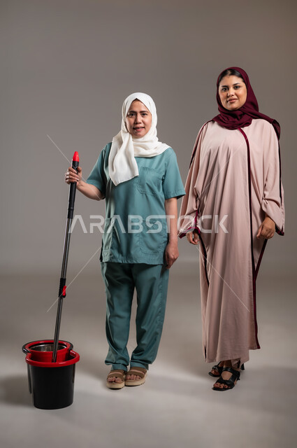 النظر للكاميرا بتعابير الابتسامة، بورتريه لإمرأة عربية خليجية سعودية ومساعدة منزلية فلبينية ترتدي اليونيفورم الخاص بها مستعدة للقيام بالعمل، ايماءات سعادة وسرور، ادوات تنظيف بلون أحمر و أسود، استقدام العماله من الخارج، الطول الكامل للجسم، خلفية رمادية