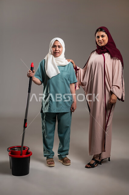 النظر للكاميرا بتعابير السعادة، بورتريه لإمرأة عربية خليجية سعودية محجبة تقف بجانب الخادمة الفلبينية بايماءات السرور، الاستعداد للقيام بالاعمال المنزلية، استقدام العماله من الخارج، خلفية رمادية