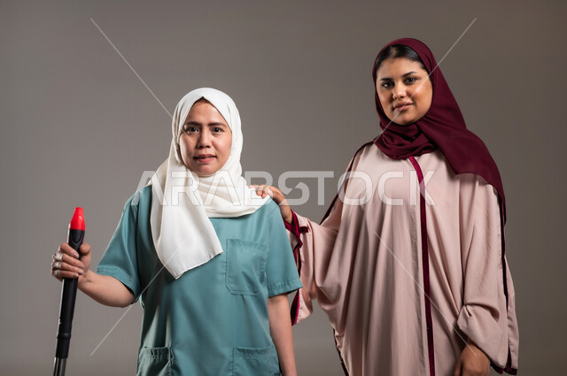 النظر للكاميرا بتعابير السعادة، بورتريه لإمرأة عربية خليجية سعودية محجبة تقف بجانب الخادمة الفلبينية بايماءات السرور، الاستعداد للقيام بالاعمال المنزلية، استقدام العماله من الخارج، خلفية رمادية