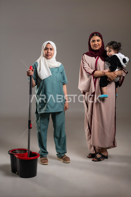 النظر للكاميرا بتعابير السعادة، بورتريه لإمرأة عربية خليجية سعودية محجبة تقف بجانب الخادمة الفلبينية بايماءات السرور، الاستعداد للقيام بالاعمال المنزلية، استقدام العماله من الخارج، الجسم بالطول الكامل، خلفية رمادية