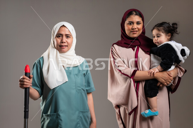 النظر للكاميرا بتعابير السعادة، بورتريه لإمرأة عربية خليجية سعودية محجبة تقف بجانب الخادمة الفلبينية بايماءات السرور، الاستعداد للقيام بالاعمال المنزلية، استقدام العماله من الخارج، خلفية رمادية