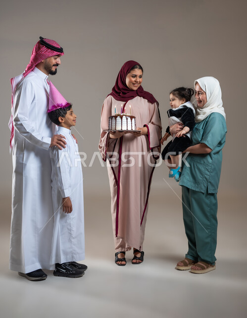 المشاركة بأعياد الميلاد، الاحتفال بأعياد الميلاد، بورتريه لعائلة عربية خليجية تحتفل بالذكرى السنوية لعاملة فلبينية، أجواء فرحة لأسرة سعودية، كيكة مزينة مع الشموع ، الاستمتاع و الترفيه بأجواء الاحتفالات, خلفية رمادية.