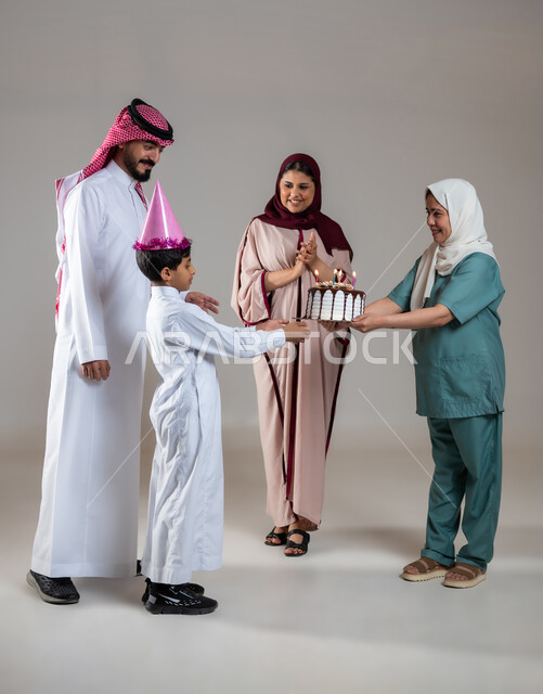 أجواء مليئة بالحب، قضاء أوقات ممتعه بالمناسبات وأعياد الميلاد, بورتريه لعائلة عربية سعودية خليجية تحتفل بالعاملة الفيلبينية وتقدم لها قالب الكيك المزين بالشموع، ايماءات وجه تدل على السعادة و السرور, الاهتمام بمشاعر الغير، الطول الكامل للجسم، خلفية رمادية