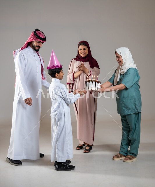 أجواء مليئة بالحب، قضاء أوقات ممتعه بالمناسبات وأعياد الميلاد, بورتريه لعائلة عربية سعودية خليجية تحتفل بالعاملة الفيلبينية وتقدم لها قالب الكيك المزين بالشموع، ايماءات وجه تدل على السعادة و السرور, الاهتمام بمشاعر الغير، الطول الكامل للجسم، خلفية رمادية
