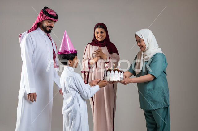 الاهتمام بمشاعر الغير، قضاء أوقات ممتعه بالمناسبات وأعياد الميلاد, بورتريه لعائلة عربية سعودية خليجية تحتفل بالعاملة الفيلبينية وتقدم لها قالب الكيك المزين بالشموع، ايماءات وجه تدل على السعادة و السرور, أجواء مليئة بالحب، خلفية رمادية