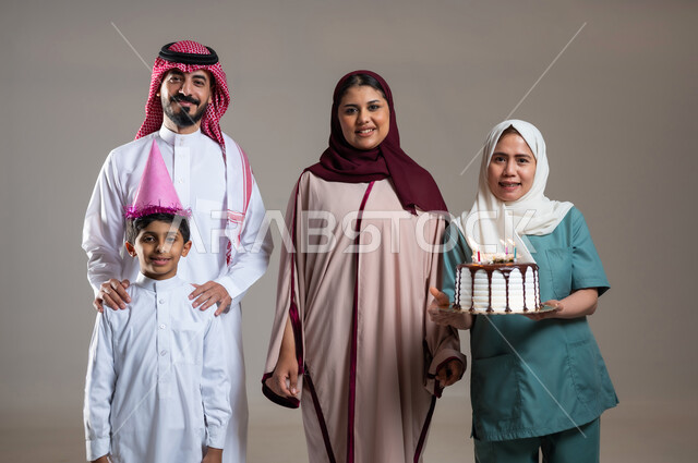 الاهتمام بمشاعر الغير، قضاء أوقات ممتعه بالمناسبات وأعياد الميلاد, بورتريه لعائلة عربية سعودية خليجية تحتفل بالعاملة الفيلبينية بالكيك المزين بالشموع، النظر الى الكاميرا بايماءات السعادة و السرور, أجواء مليئة بالحب، خلفية رمادية