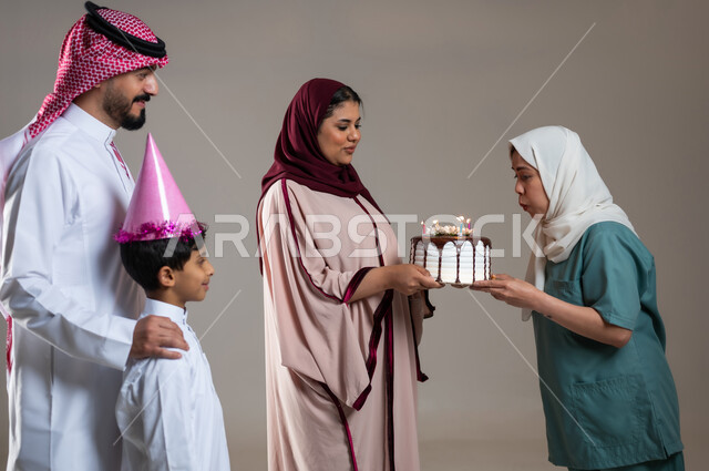 النفخ على شموع كعكة عيد الميلاد، قضاء أوقات ممتعه بالمناسبات وأعياد الميلاد, بورتريه لعائلة عربية سعودية خليجية تحتفل بالعاملة الفيلبينية بالكيك المزين بايماءات السعادة و السرور, أجواء مليئة بالحب، الاهتمام بمشاعر الغير، خلفية رمادية