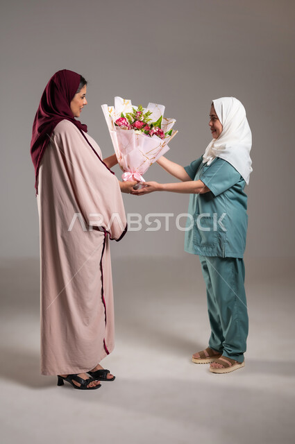 ادخال السرور الى قلوب الغير، اعياد ومفاجآت ومناسبات سعيدة، أجواء مليئة بالتقدير والاحترام، بورتريه من الجانب لإمرأة عربية سعودية خليجية تقوم بتقديم باقة من الزهور لعاملة فلبينية بايماءات السعادة و السرور, الطول الكامل للجسم، خلفية رمادية