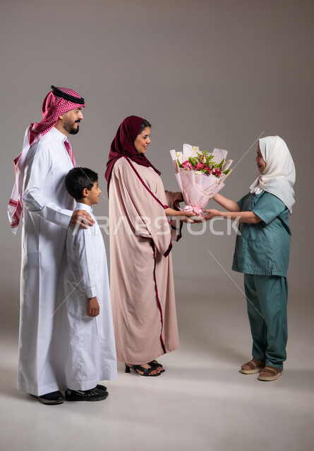 مشاركة وتقديم الهدايا بالمناسبات وأعياد الميلاد, بورتريه لعائلة عربية سعودية خليجية تقوم بتقديم باقة من الزهور لعاملة فلبينية بايماءات السعادة و السرور, أجواء مليئة بالتقدير والاحترام، الاهتمام بمشاعر الغير، الطول الكامل للجسم, خلفية رمادية