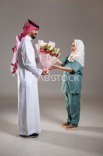 الإحسان في التعامل مع الخادمات في البيوت السعودية، القيام بالمهام المنزلية الروتينية، بورتريه لرجل عربي سعودي خليجي يقوم بتقديم باقة زهور للعاملة الفلبينية بايماءات الشكر والامتنان لجودة خدمتها وادائها، مهن ووظائف نسائية، الطول الكامل للجسم، خلفية رمادية