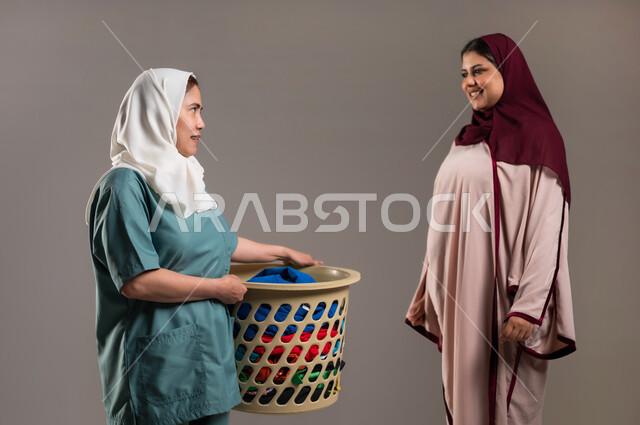 حوارات ونقاشات مدبرة المنزل مع خادمة منزلية أجنبية، بورتريه لعاملة فلبينية ترتدي اليونيفورم الخاص بالعمل وتحمل سلة الملابس، القيام بالمهام والأعمال المنزلية الروتينية، ايماءات السعادة والرضا عن جودة خدمة العاملة، خلفية رمادية