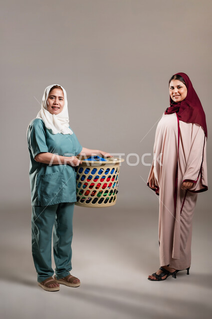 تأجير العمالة الخارجية المرخصة والمؤهلة، رضا وسعادة ربة المنزل عن اداء العاملة، المعاملة الحسنة للخادمات، بورتريه لسيدة عربية خليجية سعودية مع خادمة فلبينية تحمل سلة ملابس تنظران الى الكاميرا، الطول الكامل للجسم, خلفية رمادية