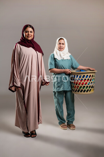 رضا وسعادة ربة المنزل عن اداء العاملة،تأجير العمالة الخارجية المرخصة والمؤهلة، المعاملة الحسنة للخادمات، بورتريه لسيدة عربية خليجية سعودية مع خادمة فلبينية تحمل سلة ملابس تنظران الى الكاميرا، الطول الكامل للجسم, خلفية رمادية